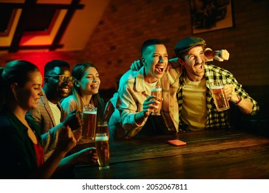 Multi-ethnic Group Of Friends Having Fun While Watching Sports Match On TV And Drinking Beer In A Pub.