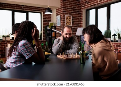 Multiethnic group of friends playing chess boardgame at home. Diverse people sitting at home, in living room, enjoying board games, snacks and beverages while having a good time together.