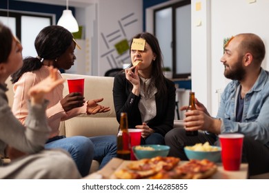 Multiethnic Group Of Friends Using Sticky Notes To Play Guess Who Game Together At Office Party After Hours. Business People Playing Charades, Eating Pizza And Drinking Beer After Work.