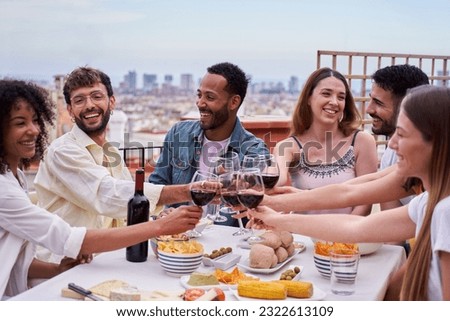 Multi-ethnic group of happy people toasting with red wine on a rooftop in the city. Friends having fun together, celebrating in a terrace during daytime, sitting at a table full of food. 