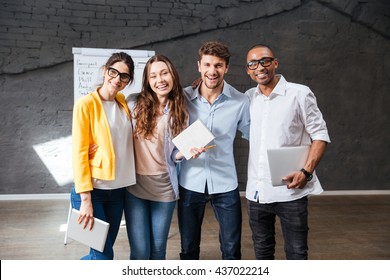 Multiethnic Group Of Happy Young Business People Holding Tablet And Laptop Standing In Office