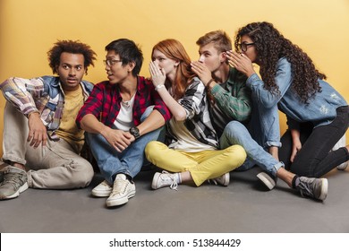 Multiethnic group of happy young friends sitting and whispering secrets to african boy over yellow background