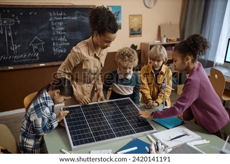 Multiethnic group of kids learning about renewable energy in school with young teacher sowing solar panel to class