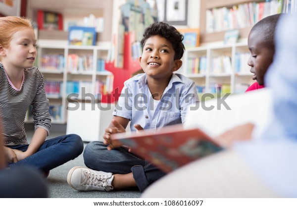 Multiethnic group of kids sitting on floor in\
circle around the teacher and listening a story. Discussion group\
of children inaa library talking to woman. Smiling hispanic boy in\
elementary school.
