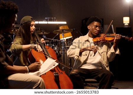 Multiethnic group of musicians playing string instruments in music studio, Black boy performing on violin while Latin girl playing cello, another man holding sheet music