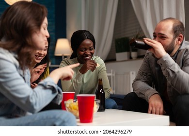 Multi-ethnic group of people enjoying spending time together laughing during wekeend party sitting on sofa in living room. Friends drinking beer, eating snack. Hanging out concept