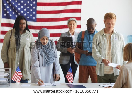 Multi-ethnic group of people registering at polling station decorated with American flags on election day, focus on Arab woman signing ballot form in foreground, copy space