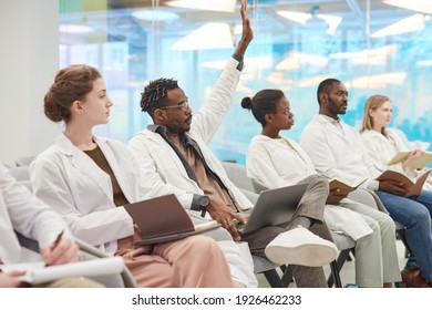 Multi-ethnic group of people wearing lab coats sitting in row in audience at medical seminar, focus on African American man raising hand to ask question, copy space - Shutterstock ID 1926462233