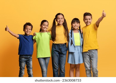 Multiethnic group of school aged kids in casual outfit cheerfully smiling at camera, embracing and showing thumb up posing isolated on yellow background. Happy childhood, kids, friendship concept - Powered by Shutterstock