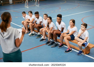 Multi-ethnic Group Of School Kids Listening To Their Sports Teacher During Physical Education Class At School Gym.