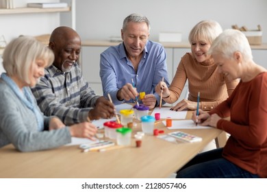 Multiethnic group of senior people having painting activity at sanatorium, cheerful elderly men and women in casual outfits sitting around table in living room, drawing and having conversation