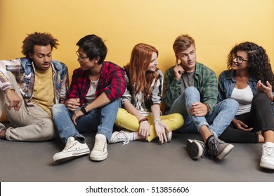 Multiethnic Group Of Smiling Young Friends Sitting And Talking Over Yellow Background