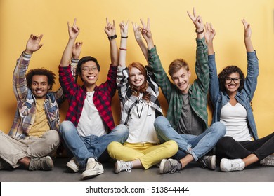 Multiethnic Group Of Smiling Young People Celebrating Success And Showing Peace Sign With Raised Hands Over Yellow Background