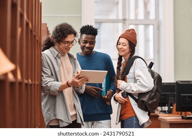 Multiethnic group of three cheerful young people standing by shelves in college library and using digital tablet copy space - Powered by Shutterstock