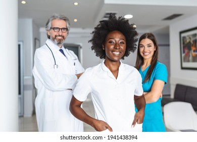 A Multi-ethnic Group Of Three Doctors And Nurses Standing In A Hospital Corridor, Wearing Scrubs And Coats. The Team Of Healthcare Workers Are Staring At The Camera And Smiling