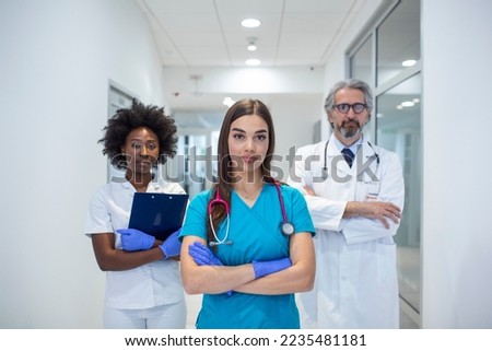 A multi-ethnic group of three doctors and nurses standing in a hospital corridor, wearing scrubs and coats. The team of healthcare workers are staring at the camera and smiling