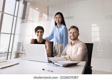 Multiethnic group of three positive professionals posing for portrait at workplace with laptop. Diverse business team meeting for teamwork at large office table, looking at camera, smiling - Powered by Shutterstock