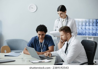 Multiethnic group of three young doctors using laptop together in clinic with X rays in background, copy space