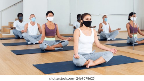 Multiethnic group of young adults in face masks for viral protection sitting in lotus position practicing meditation at yoga class - Powered by Shutterstock