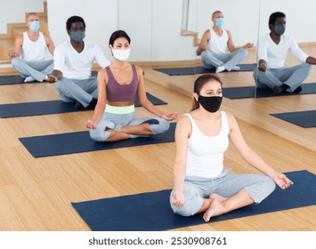 Multiethnic group of young adults in face masks for viral protection sitting in lotus position practicing meditation at yoga class - Powered by Shutterstock