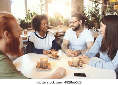 Multiethnic Group Of Young Ambitious People Sitting At Round Table At Coworking Space, Working Together On New Business Strategy Of Their Small Company, Discussing Plans And Developing Solutions