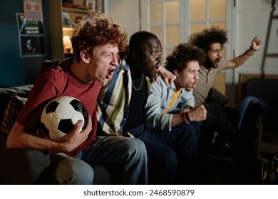 Multi-ethnic group of young male soccer fans sitting on couch in living room watching match on TV - Powered by Shutterstock