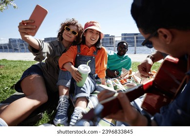 Multiethnic group of young people taking selfies outdoors enjoying picnic on grass in sunlight, copy space - Powered by Shutterstock