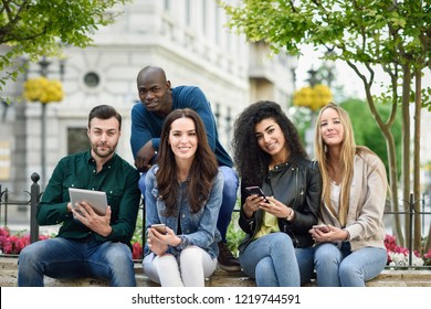 Multi-ethnic group of young people using smartphone and tablet computers outdoors in urban background. Women and men smiling and laughing in the street wearing casual clothes.