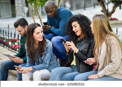 Multi-ethnic group of young people using smartphone and tablet computers outdoors in urban background. Women and men smiling and laughing in the street wearing casual clothes.