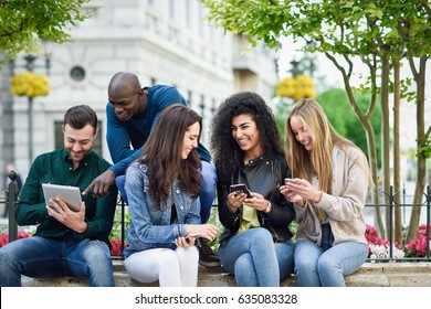 Multi-ethnic group of young people using smartphone and tablet computers outdoors in urban background. Women and men smiling and laughing in the street wearing casual clothes.