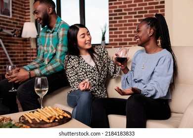 Multiethnic joyful women at wine party talking while sitting on sofa. Happy smiling diverse friends sitting in living room while enjoying alcoholic beverages together while celebrating birthday