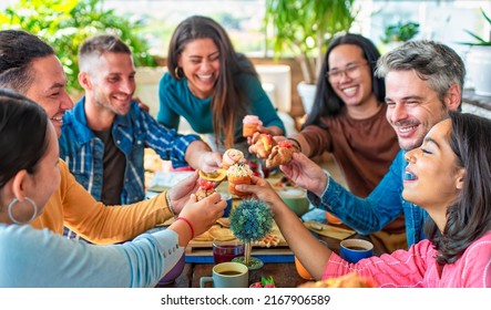 Multiethnic Large Group Of Friends Sitting On Cafe Table Restaurant Eating A Muffin Making Faces. Diverse People Celerating Sweet Breakfast Together Enjoying Happy Holidays. Lifestyle And Joy Concept