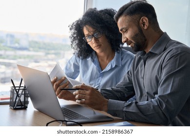 Multiethnic male indian mentor and female African American intern sitting at desk with laptop doing paperwork together discussing project financial report. Corporate business collaboration concept.