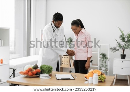 Multiethnic man in doctor's coat finding out waist length of female patient with measuring tape in consulting room. Efficient dietitian calculating body mass index for assessing healthy weight.