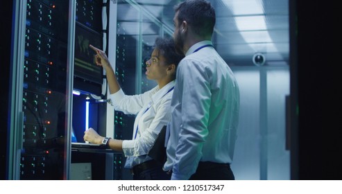 Multiethnic man and woman in white shirts using laptop and tablet while working in server room of modern data center