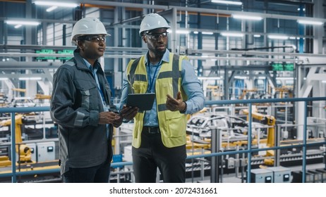 Multiethnic Manager And African American Car Factory Engineer In Uniform Using Tablet Computer. Automotive Industry 4.0 Manufacture Employees Discuss Work On Vehicle Assembly Plant.