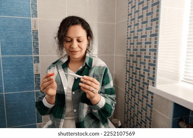 Multi-ethnic middle-aged woman in green checkered shirt, sitting on the toilet bowl in the home bathroom, taking pregnancy test, awaiting the result. People, lifestyle, maternity and pregnancy concept - Powered by Shutterstock