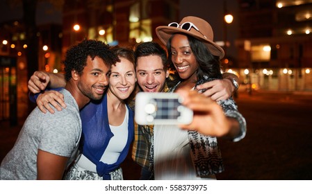 Multi-ethnic millenial group of friends taking a selfie photo with mobile phone on rooftop terrasse using flash at night time