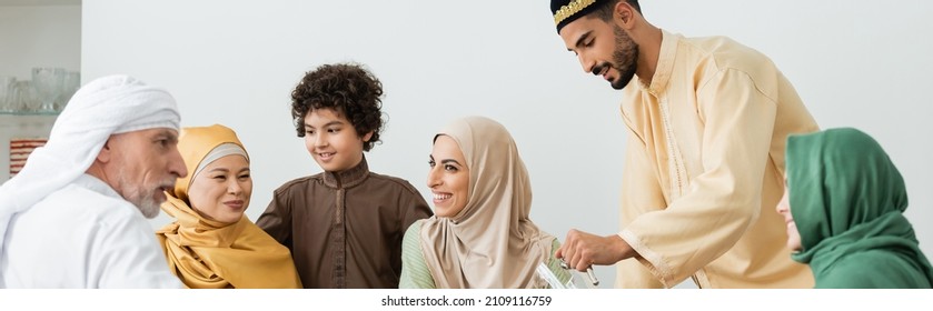 multiethnic muslim family smiling near arabian man with jug at home, banner