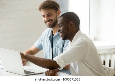 Multiethnic office workers laughing watching funny videos at laptop, African American guy showing something to Caucasian colleague. Diverse team at casual break, smiling, having fun, good relations
