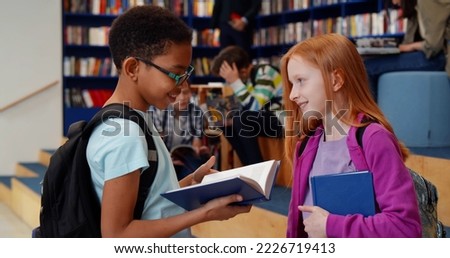 Multiethnic schoolchildren reading book and talking in library. Side view of african schoolboy and caucasian schoolgirl doing homework or preparing for test together in library