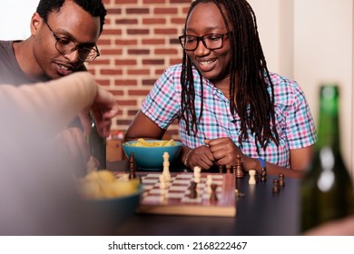 Multiethnic smart friends playing a game of chess while sitting at table in living room. Happy people relaxing with snacks and beverages while enjoying strategic boardgames.