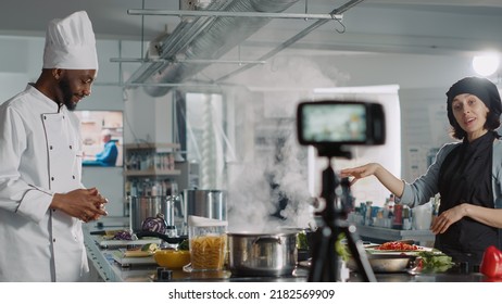 Multiethnic Team Of Chefs Hosting Online Cooking Show, Filming Video On Camera In Restaurant Kitchen. Man And Woman Doing Gastronomic Culinary Class, Recording Food Recipe Preparations.