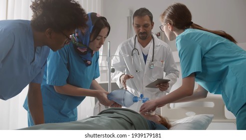 Multiethnic Team Of Doctors Putting Oxygen Mask On Patient In Hospital. Indian Doctor Supervising And Teaching Interns Resuscitating Patient Using Oxygen Bag In Hospital