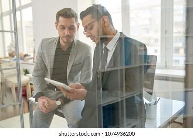 Multi-ethnic team of financial managers gathered together at spacious office and analyzing statistic data with help of digital tablet, view through glass wall