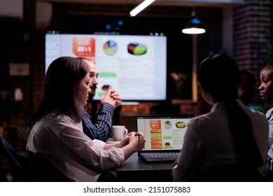 Multiethnic team in night business meeting doing overtime having sales statistics presentation sitting at desk. Workaholic colleagues doing teamwork in late conference looking at statistics on screen.