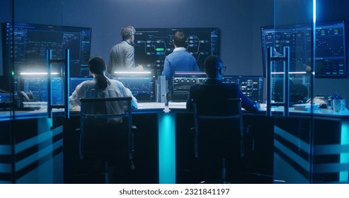 Multiethnic team of IT technical specialists and software programmers work with data server, analysis charts and blockchain network on computers and big digital screens in monitoring office at night.