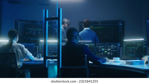 Multiethnic team of IT technical specialists and software programmers work with data server, analysis charts and blockchain network on computers and big digital screens in monitoring office at night.