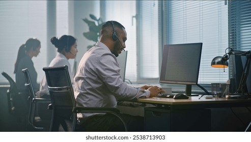 Multiethnic team of technical support operators working in modern call center office. African American helpdesk specialist talks on headset, uses computer, looks at camera providing customer service.