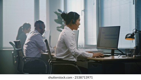 Multiethnic team of technical support operators working in modern call center office. Asian female helpdesk specialist talks on headset, uses computer, looks at camera providing customer service.